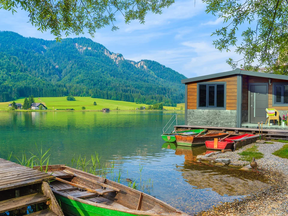 Compact modular cabin right on a mountain lake shore clad in dark wood-toned fibre cement siding planks